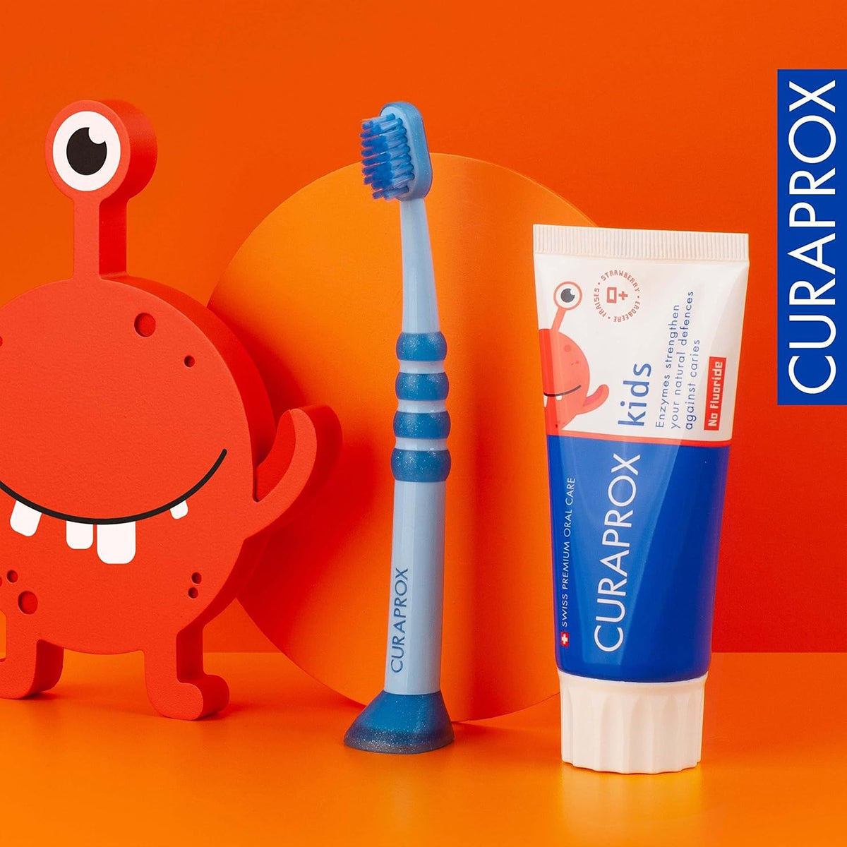 Curaprox Kids tandpasta aardbei naast blauwe tandenborstel en oranje monstertje