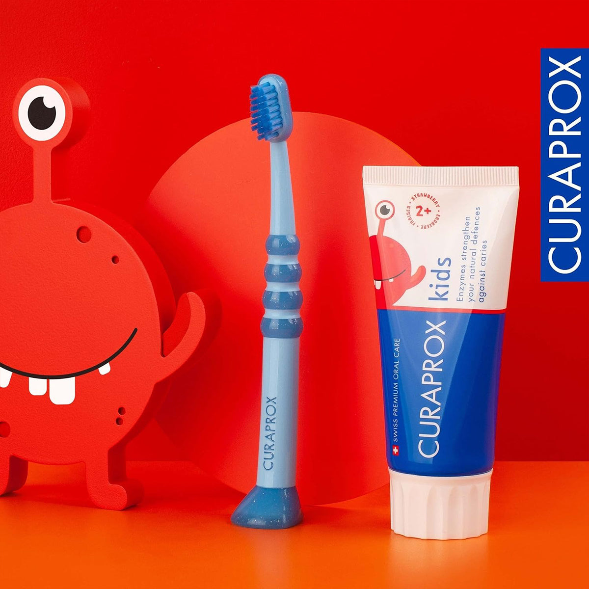 Curaprox Kinder tandpasta aardbei 2+ jaar naast blauwe kindertandenborstel en monstertje
