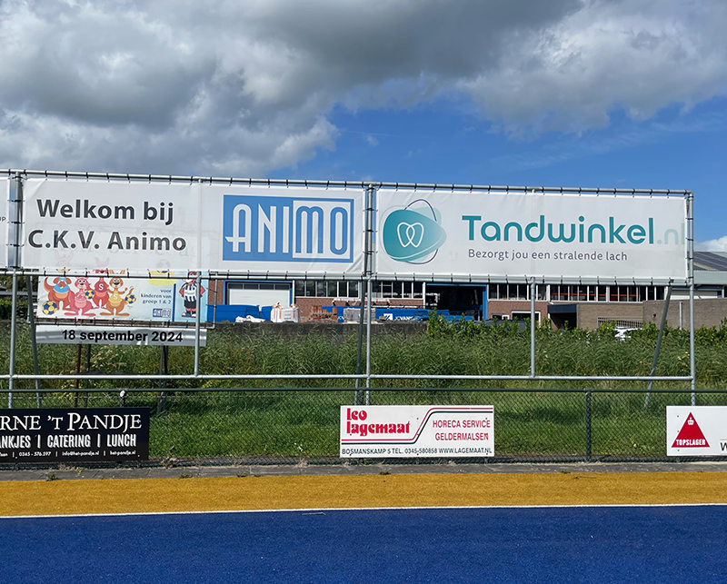 Reclamebord Tandwinkel.nl met tekst 'Bezorgt jou een stralende lach' bij sportveld